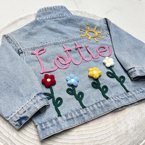 May include: Light blue denim jacket with the name "Lottie" in pink script, a yellow sun, and crocheted flowers in red, blue, yellow, and white. The jacket has a classic collar and button closure.