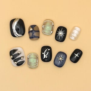 May include: A collection of ten artificial nails in various designs. The nails feature a range of colors, including black, white, blue, and green, with metallic accents and geometric patterns. Some nails have a crescent moon, sun, or star design.
