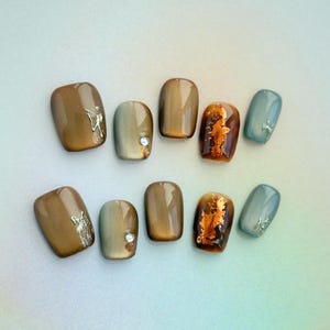 May include: A set of ten press-on nails in various shades of brown, gold, and blue. Some nails feature silver accents, a small gem, or a gold foil design. The nails are arranged on a gradient background.