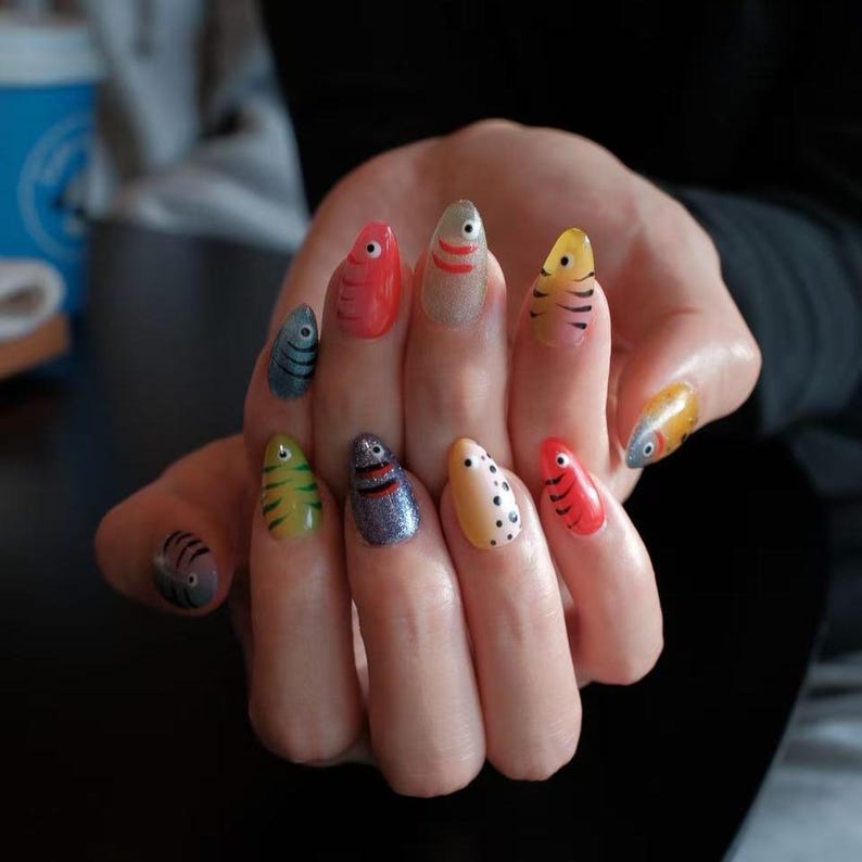 May include: A hand displaying a set of painted fingernails with a fish design. Each nail features a different color and pattern, including red, yellow, blue, and glittery silver, with details like stripes, dots, and eye designs. The nails are almond-shaped.