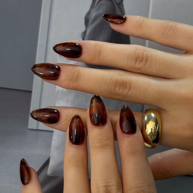 Pu&ograve; includere: Primo piano di una mano con unghie lunghe a mandorla dipinte con un motivo marrone scuro, tartarugato. Un anello d'oro &egrave; indossato su un dito. Le unghie hanno una finitura lucida.
