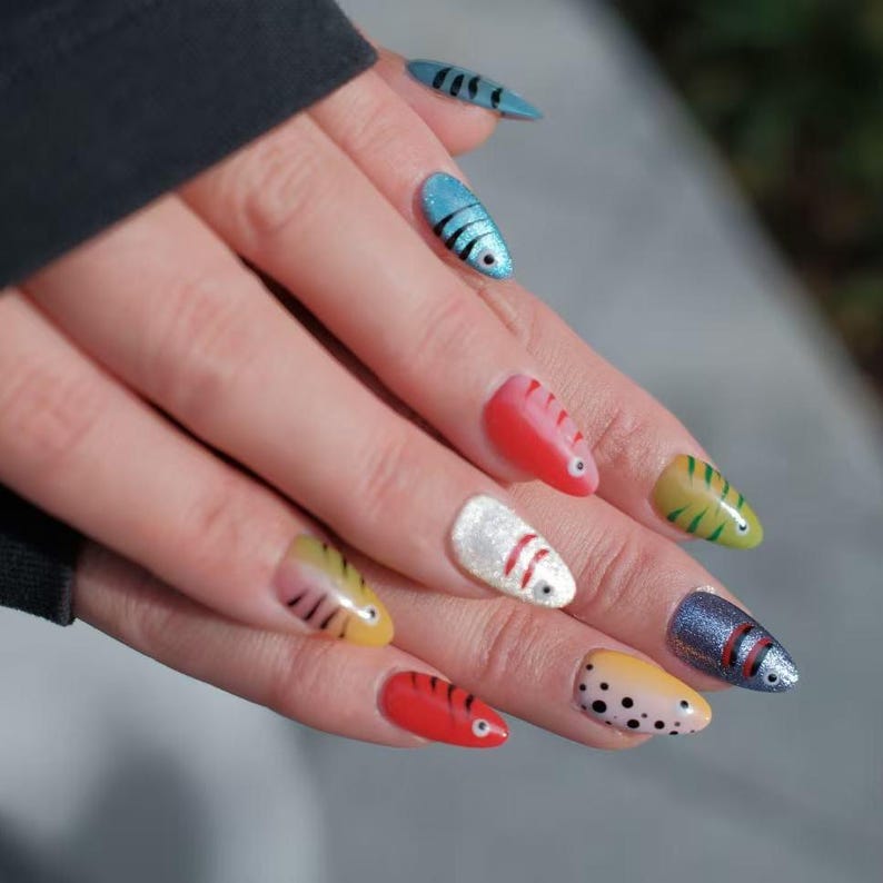 May include: A hand with painted fingernails in various colors and designs. The nails are shaped like fish, with details including stripes, dots, and eye accents. Colors include red, blue, yellow, and white.