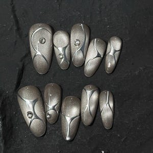 May include: A set of ten oval-shaped, press-on nails. Each nail is a shimmering taupe color with silver geometric designs and a small rhinestone accent. The nails are arranged on a dark surface.