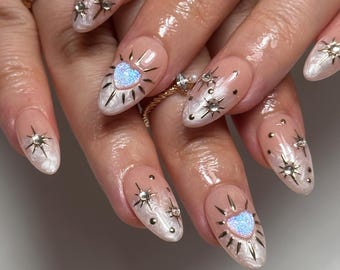 May include: Close-up of a manicure featuring almond-shaped nails with a pearlescent base. Each nail is adorned with star-shaped embellishments and a heart-shaped design with a glittery center. A gold ring with small stones is also visible.