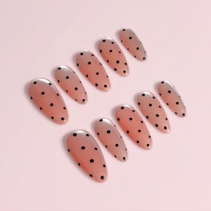May include: A set of ten almond-shaped false nails. Each nail is a nude color with small black polka dots. The nails are arranged on a light pink surface.