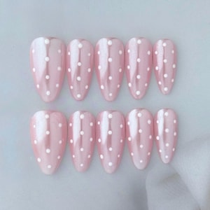 May include: A set of ten pink, almond-shaped false nails. Each nail has a pearlescent finish and is decorated with evenly spaced white polka dots. The nails are arranged in two rows of five.