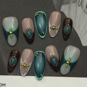 Può includere: Un set di dieci unghie finte a forma di mandorla. Le unghie presentano vari design, tra cui colori pieni, motivi bicolore e abbellimenti come perline dorate e pietre color turchese. La tavolozza dei colori include tonalità di verde, marrone, beige e bianco.