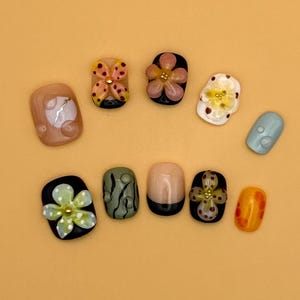 May include: A collection of ten artificial nails in various designs. The nails feature floral and abstract patterns, with colors including black, white, orange, and green. Some nails have 3D embellishments.