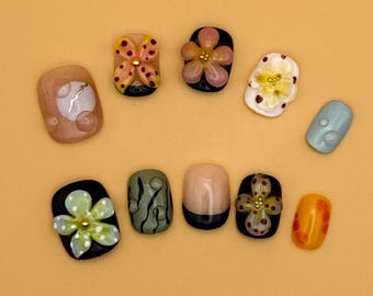 May include: A collection of ten artificial nails in various designs. The nails feature floral and abstract patterns, with colors including black, white, orange, and green. Some nails have 3D embellishments.