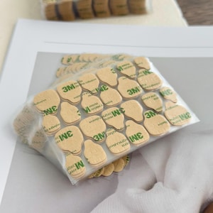 May include: A clear plastic package containing multiple beige, irregularly shaped adhesive pads. Each pad has the green text "3M" and "Adhesive" printed on it. The pads are stacked on a white surface.
