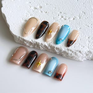 May include: A collection of ten artificial nails in various designs. The nails feature colors like tan, brown, and light blue, with accents including gold star and silver bead embellishments. Some nails have a tortoiseshell pattern or a metallic silver outline.