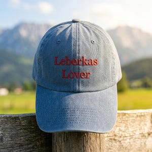 Könnte beinhalten: Eine blaue Baseballkappe aus Denim mit dem rot gestickten Text "Leberkas Lover". Die Kappe wird im Freien gezeigt, ruhend auf einem Holzzaun, mit einem verschwommenen Hintergrund aus Bergen und Grün.