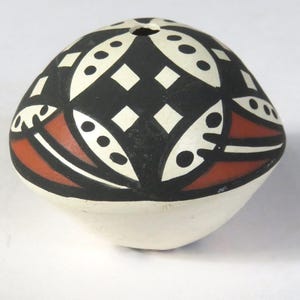 May include: A hand-painted, ceramic, egg-shaped vessel. The top is decorated with a black and white geometric pattern, with red accents. The bottom is white. The vessel has a small hole at the top. This decorative piece is likely a small pot or container.