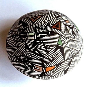 May include: A round, ceramic vessel with a black and white geometric design. The pattern features thin, parallel lines and triangular shapes in shades of brown, green, and orange. The vessel is set against a white background.