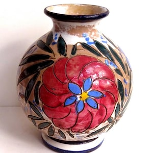 May include: A hand-painted ceramic vase with a round body and a narrow neck. The vase features a large red flower with blue and yellow accents, surrounded by green leaves. The background is tan and white, with a blue rim.