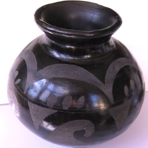 May include: A black ceramic vase with a rounded body and a flared rim. The vase features a carved, light-colored design of swirling patterns and shapes. The pottery has a glossy finish and is set against a white background.