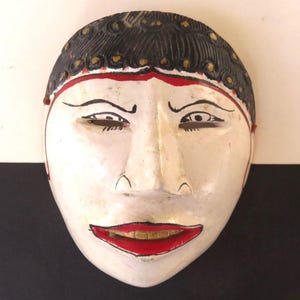 May include: A white wooden mask with black hair and red accents. The mask has a neutral expression with red lips and is set against a black and white background. The mask is likely used for theatrical or decorative purposes.