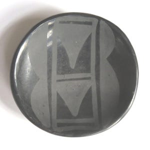 May include: A round, black ceramic bowl with a matte finish. The bowl features a symmetrical, geometric design in a lighter shade of gray, consisting of vertical lines and inverted triangles. The design is centered on the bowl's surface.