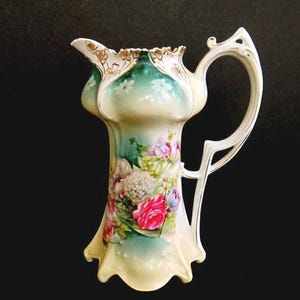 May include: An antique porcelain pitcher with a floral design. The pitcher features a white handle and a flared base. The body is painted with pink, purple, and white flowers against a green and cream background. Gold trim accents the rim.