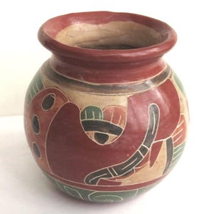 May include: A small, round, hand-painted ceramic pot with a wide opening. The pot is primarily red with tan, green, and black decorative elements. The rim is a darker shade of red. The design includes abstract shapes and patterns.