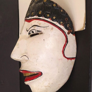 May include: A white wooden mask with black hair, gold accents, and red details. The mask has a side profile with a red outline, black eyebrows, and red lips with gold teeth. The mask is likely a decorative or theatrical piece.