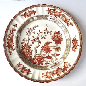 May include: A decorative white plate with a scalloped edge and a red border. The plate features a floral design in shades of red, orange, and brown, with a detailed pattern around the center.