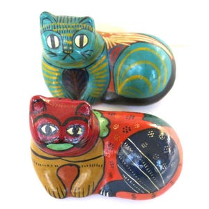 May include: Two hand-painted cat figurines. The top cat is teal with yellow stripes and accents. The bottom cat is red with orange and blue details. Both have large eyes and are in a resting position. These decorative cats are likely made of wood.