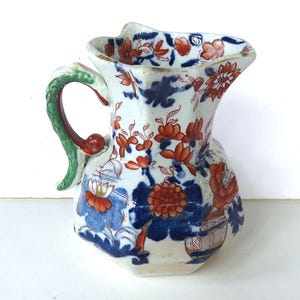 May include: Antique ceramic pitcher with a white base, adorned with intricate floral patterns in shades of blue and orange. The handle is green with red accents. The pitcher has a unique, scalloped rim.