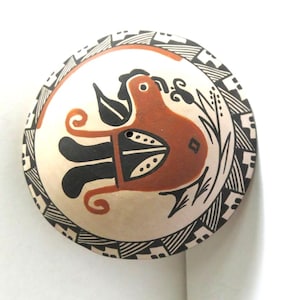 May include: Round, hand-painted ceramic bowl with a bird design. The bird is in shades of brown and black, with a cream-colored background. The rim features a black and white geometric pattern. The bowl is likely a decorative piece.