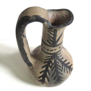 May include: A small, antique-style pottery pitcher with a rounded body and a curved handle. The pitcher is tan with black painted details, including a handle, rim, and a stylized leaf pattern. The pitcher has a spout for pouring.