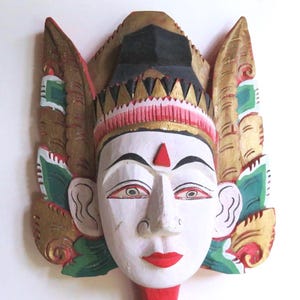 May include: Hand-carved wooden mask with a white face, red lips, and a red dot on the forehead. The mask features gold, green, and red accents, with a black and gold headdress and ornate wings. A decorative piece.