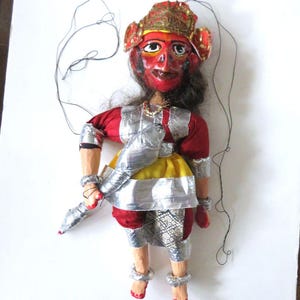 May include: A handmade puppet with a red face, wearing a red and silver outfit with a yellow skirt. The puppet has black hair and is adorned with a gold headdress. The puppet is holding a silver object and has strings attached for manipulation.