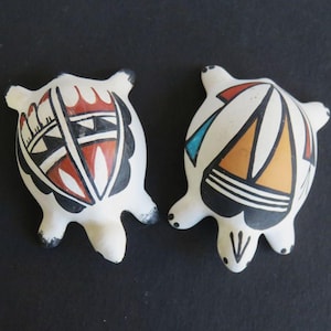 May include: Two small, white ceramic turtle figurines with painted geometric designs in red, black, orange, and blue. The turtles have black feet and decorative patterns on their shells.
