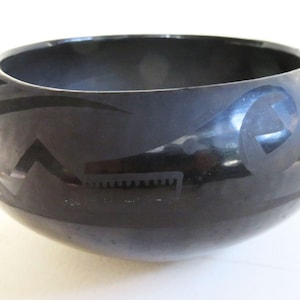 May include: A polished black ceramic bowl featuring a rounded silhouette and a wide opening. The exterior surface displays a matte black geometric pattern that contrasts against the glossy finish of the bowl's body.