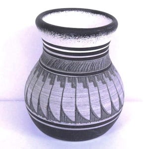 May include: A decorative ceramic vase with a rounded body and a narrow neck. The vase features a black base and rim, with a white and black striped section near the top. The body is decorated with a repeating geometric pattern in shades of gray.