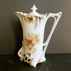 May include: A white porcelain pitcher with a lid, featuring a floral design in orange, red, and green. The pitcher has a gold trim and a unique handle. The base has an openwork design with gold accents. A decorative piece.