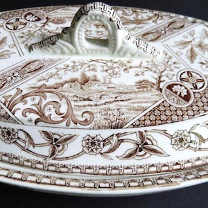 May include: A vintage ceramic serving dish lid with a brown and white transferware design. The lid features a landscape scene with a bridge and buildings, surrounded by floral and geometric patterns. The handle is arched and detailed.