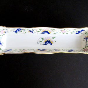 May include: A rectangular white porcelain dish with a gold rim and floral design. The dish features a repeating pattern of blue, pink, and yellow flowers and leaves. The dish is approximately 10 inches long and 4 inches wide.