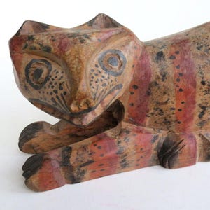 May include: Wooden cat sculpture with a hand-painted design. The cat is lying down, with a detailed face and body. The sculpture features red, black, and brown colors, with a rustic, folk art style.