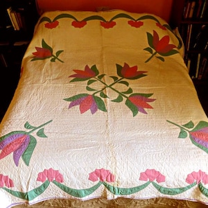 May include: Cream-colored quilt with a floral design. The quilt features red, pink, green, and purple tulip-like flowers. The flowers are arranged in a symmetrical pattern across the quilt's surface. The quilt is on a bed.