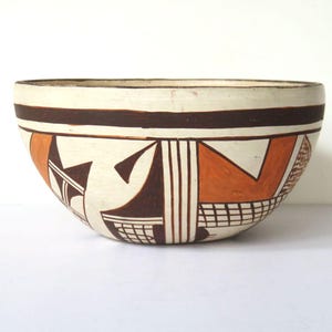 May include: A ceramic bowl with a cream-colored exterior and a brown band near the rim. The bowl features geometric designs in brown and orange, including triangles, lines, and a grid pattern. The bowl is likely handmade.