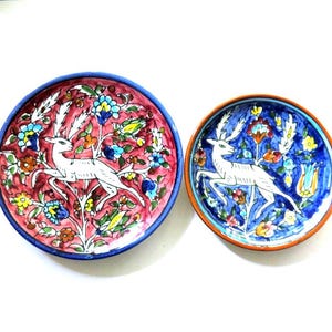 May include: Two decorative ceramic plates with hand-painted designs. One plate features a red background with a white deer and floral patterns, while the other has a blue background with a similar deer and floral motif. Both plates have a colorful border.