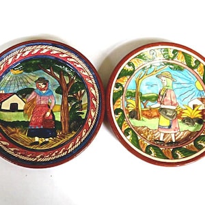 May include: Two round, hand-painted ceramic plates. Each plate features a central scene of a person in traditional attire, set within a landscape with a tree and a house. The plates have a decorative border with red, blue, and green accents.