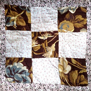 May include: A patchwork quilt with a nine-square design. The squares alternate between floral patterns in brown, gold, and blue on a dark background and white squares with small red cross patterns. The border features a floral vine pattern.