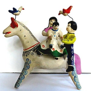 May include: A handcrafted, off-white ceramic horse figurine with red, black, and blue painted details. The horse carries three figures, and two birds perch above. The figures are dressed in colorful clothing. A unique folk art piece.