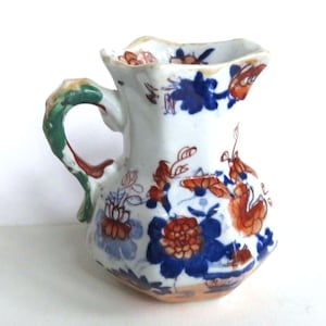 May include: A small, antique white ceramic pitcher with a scalloped rim and a green and red handle. The pitcher is decorated with blue and orange floral patterns. The base has a light brown color.