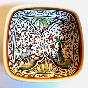 May include: Square ceramic dish with a hand-painted design. The interior features a whimsical scene with a gray donkey, floral elements, and a tower. The rim has a decorative border with orange, brown, and white accents. A unique decorative piece.