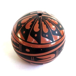 May include: A round, ceramic decorative object with a hole at the top. The object is painted with a black and reddish-brown geometric pattern. The design features curved lines, shapes, and dots. The background is white.
