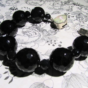 May include: A black onyx beaded bracelet with a silver clasp featuring a mother-of-pearl inlay. The bracelet is on a white floral patterned background.