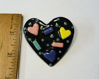 May include: A black heart-shaped pin with a glossy finish, decorated with small teal dots and raised shapes in pink, yellow, and blue. A wooden ruler is positioned to the left, showing the heart measures approximately 2 inches in height.